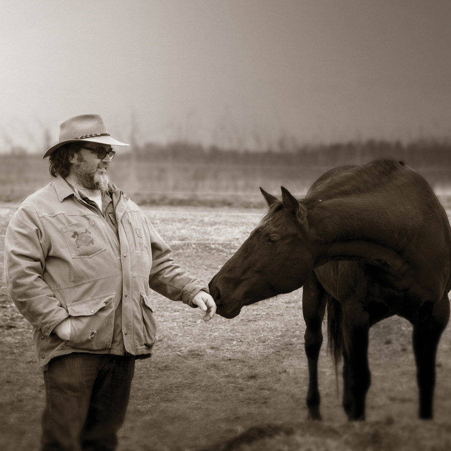 Sepia photograph: Fred, in a wide-brimmed hat, stands in a winter pasture holding out his hand to Juniper, a dark bay horse, who stretches her nose towards it.