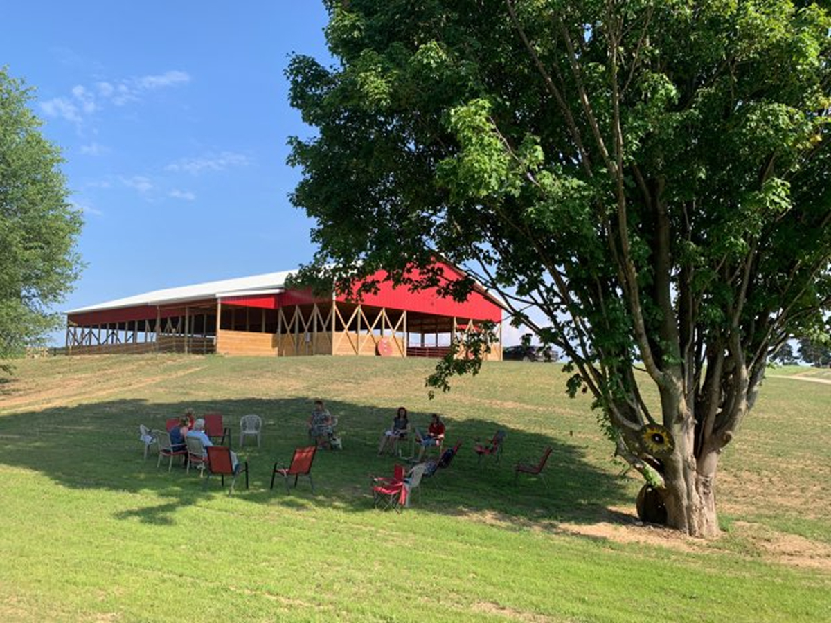 The red covered arena on the rise at Red Horse, with people sitting in a circle of chairs on the lawn below it under a maple tree.