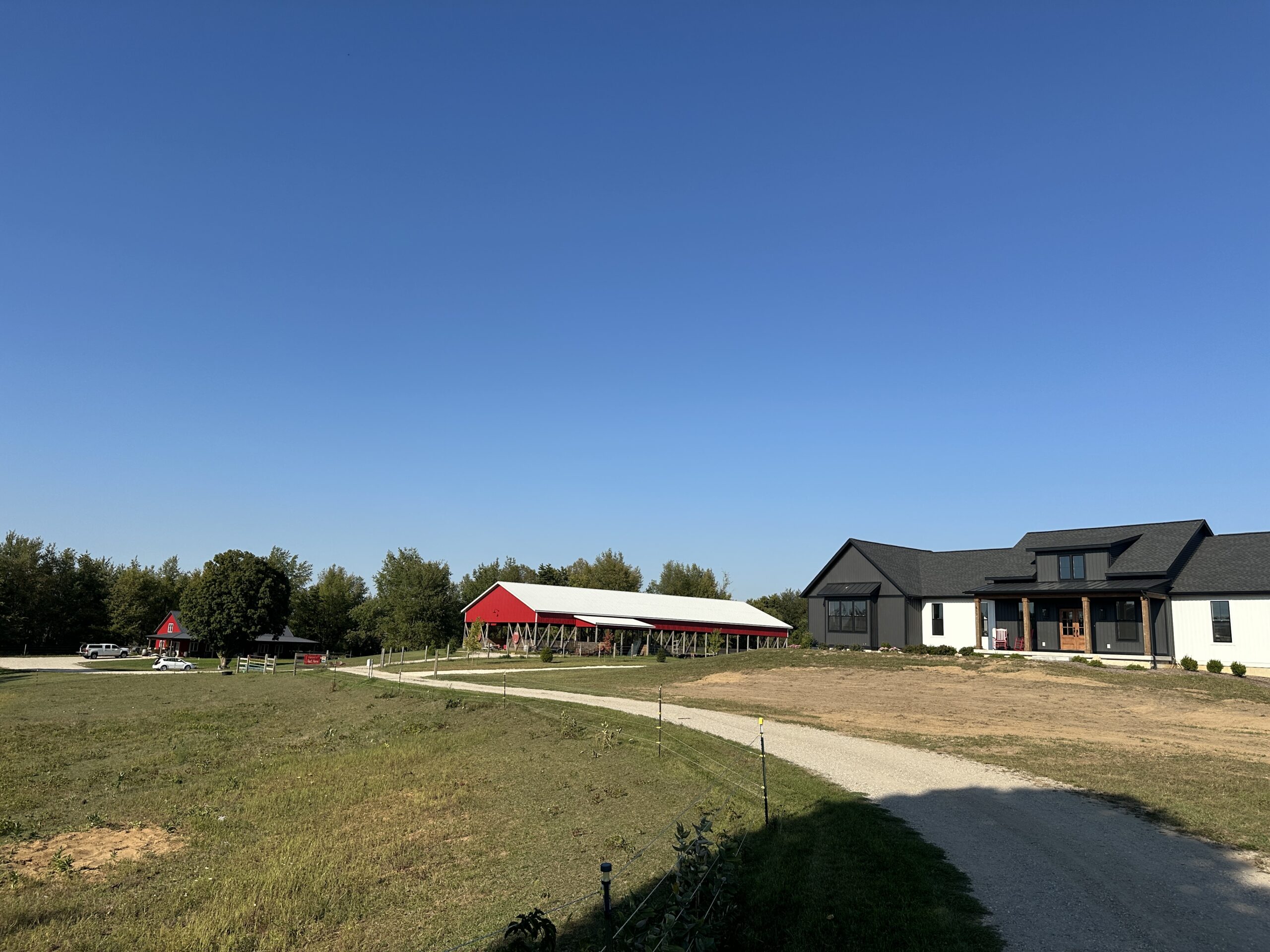 The farm seen from the gravel drive: the red barn and its open-sided pavilion on one side, the house on the other, mown grass and open sky between them.